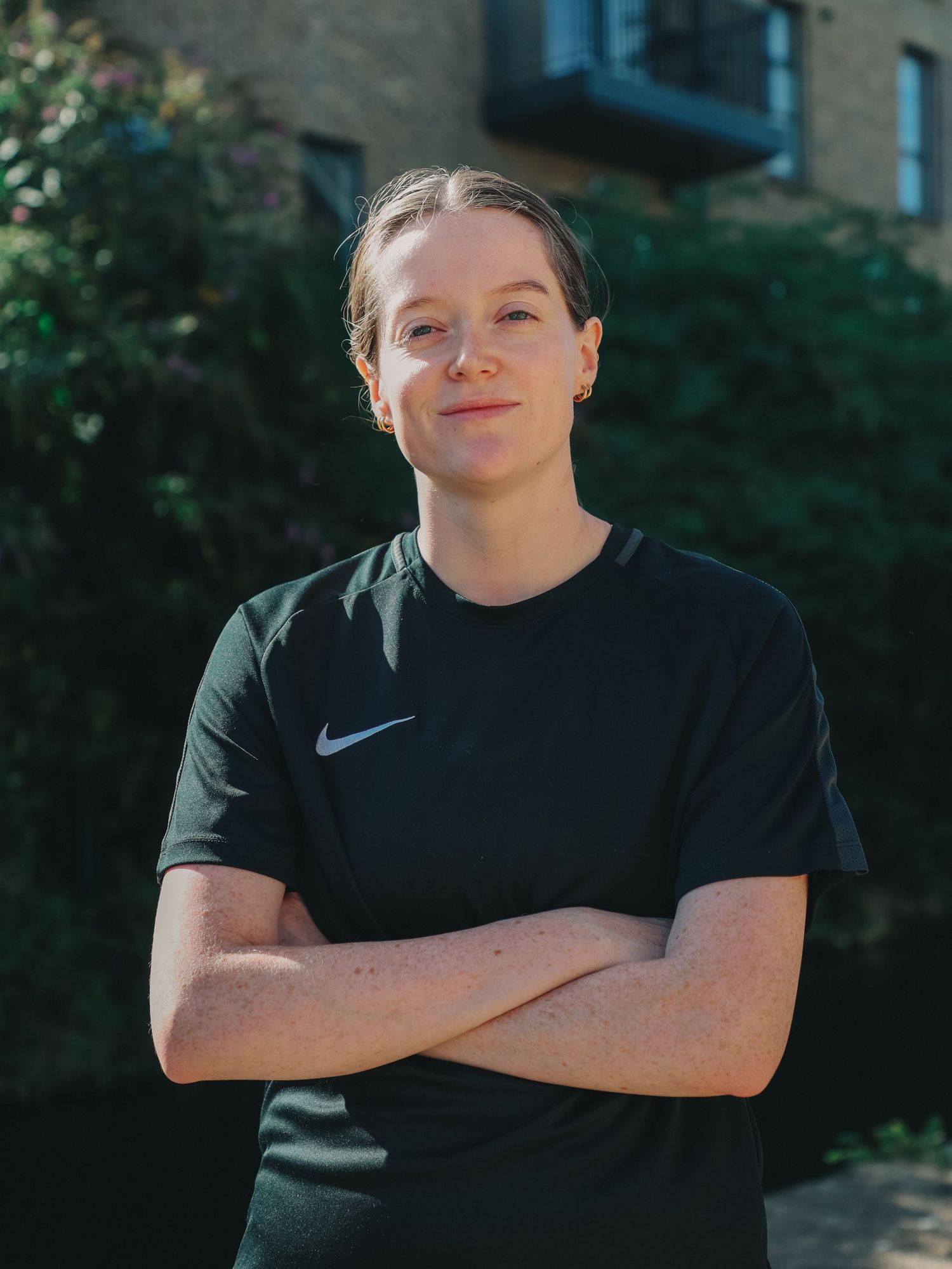 A young woman in a black Nike t-shirt stands with arms crossed, smiling outdoors against a green leafy background.