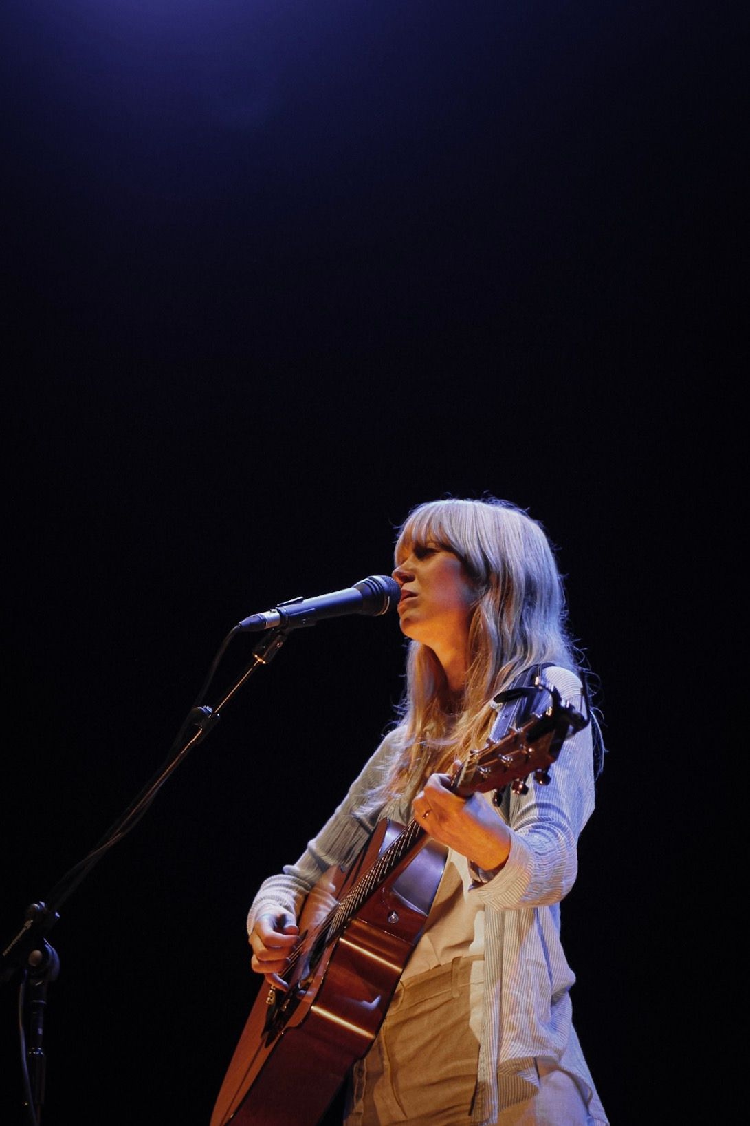 Musician in white playing acoustic guitar on darkened stage.