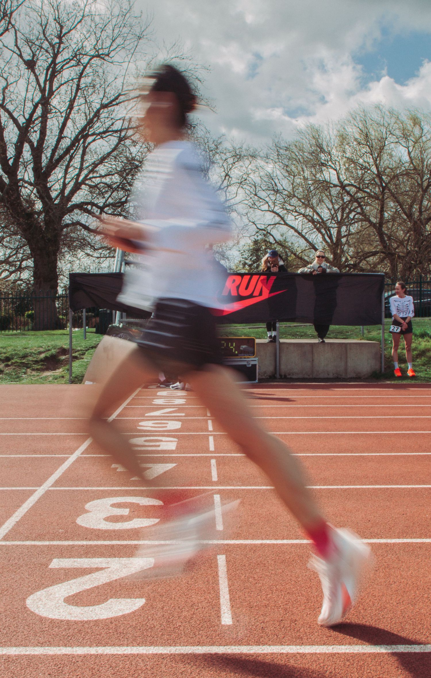 A blurred runner sprints past lane markings on a red Nike-branded athletic track outdoors.