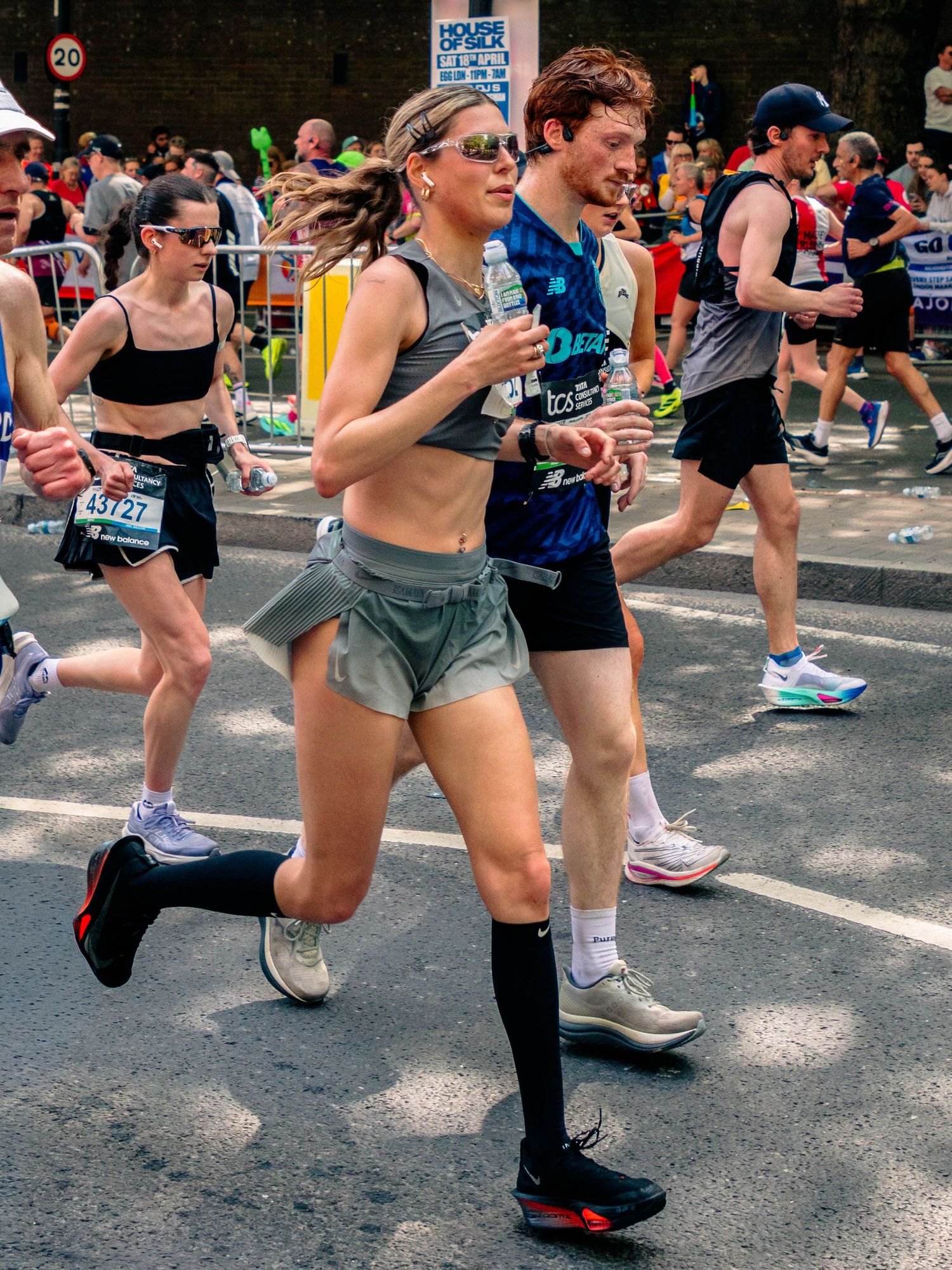 Runners competing in a city marathon, woman in gray shorts and black knee socks drinking water while racing on urban street.