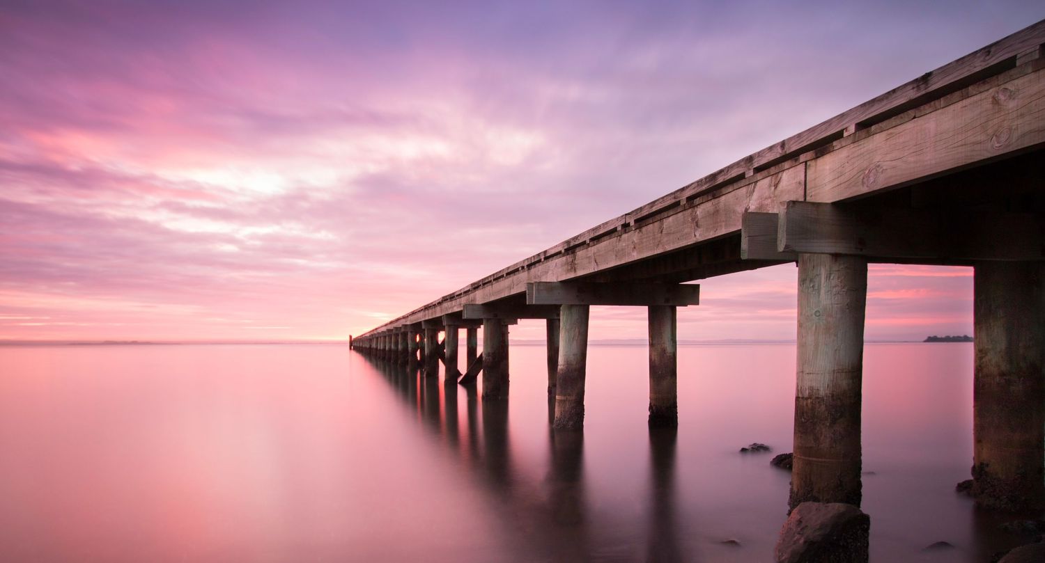 Long wooden pier stretches into calm waters under a dramatic purple sunset sky.