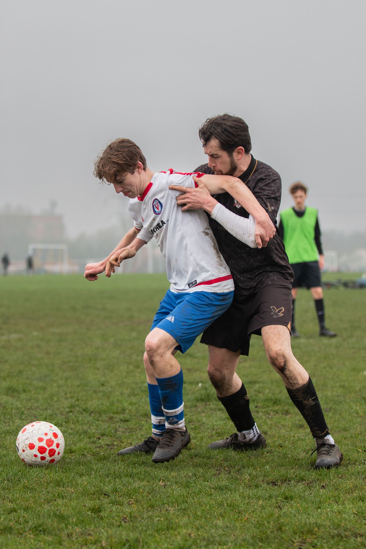 Two soccer players battle for possession of a red and white ball on a grassy field during a match.