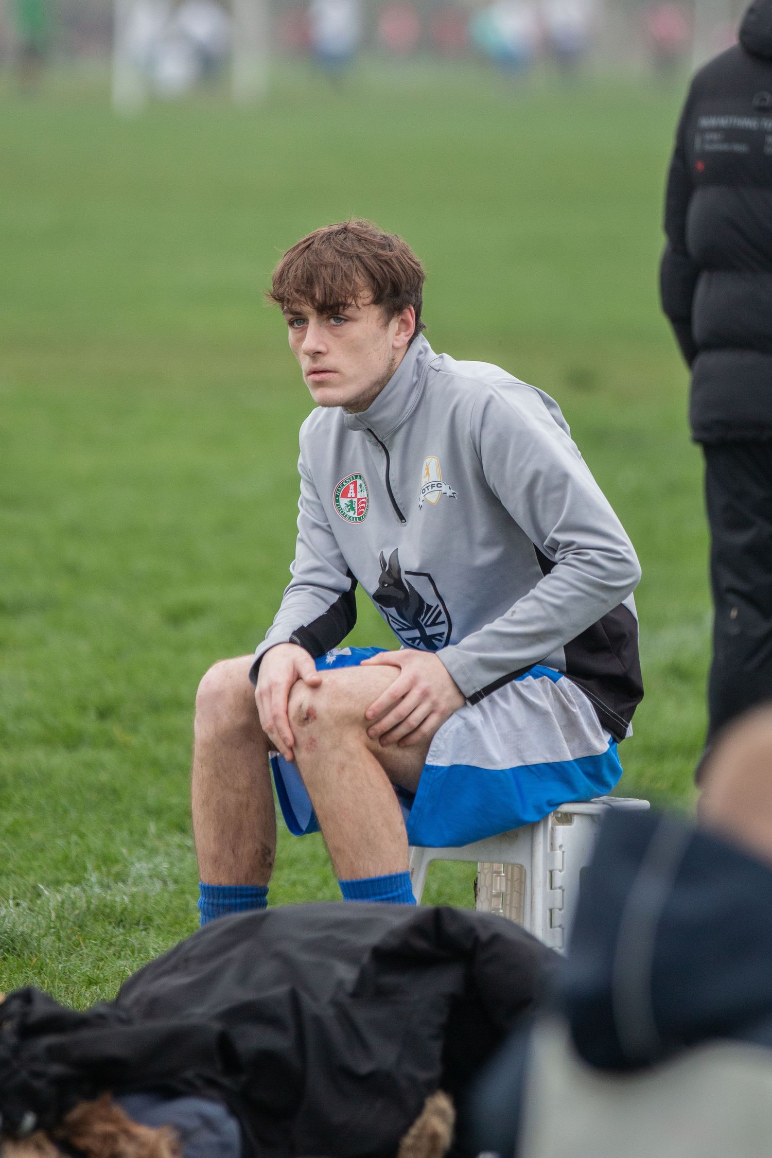 Young soccer player in gray training top and blue shorts sits on bench during outdoor practice session.