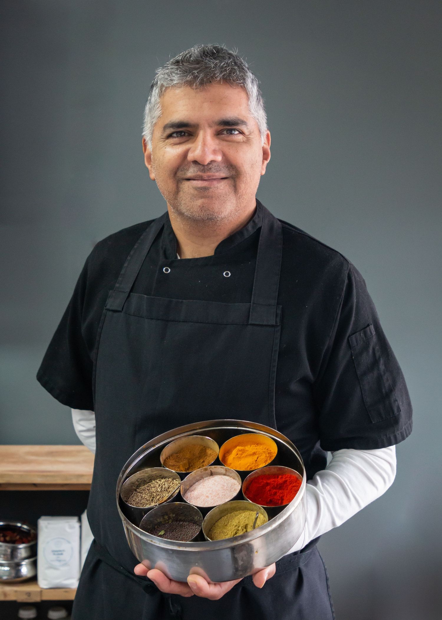 Chef of Kari Club in black uniform displays spice tins filled with colourful seasonings against grey background.