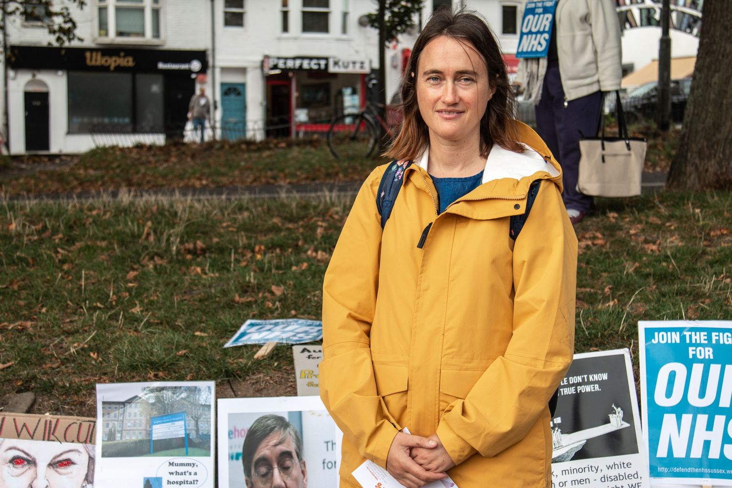 Person wearing a bright yellow raincoat stands among protest signs at a rally to save the NHS.