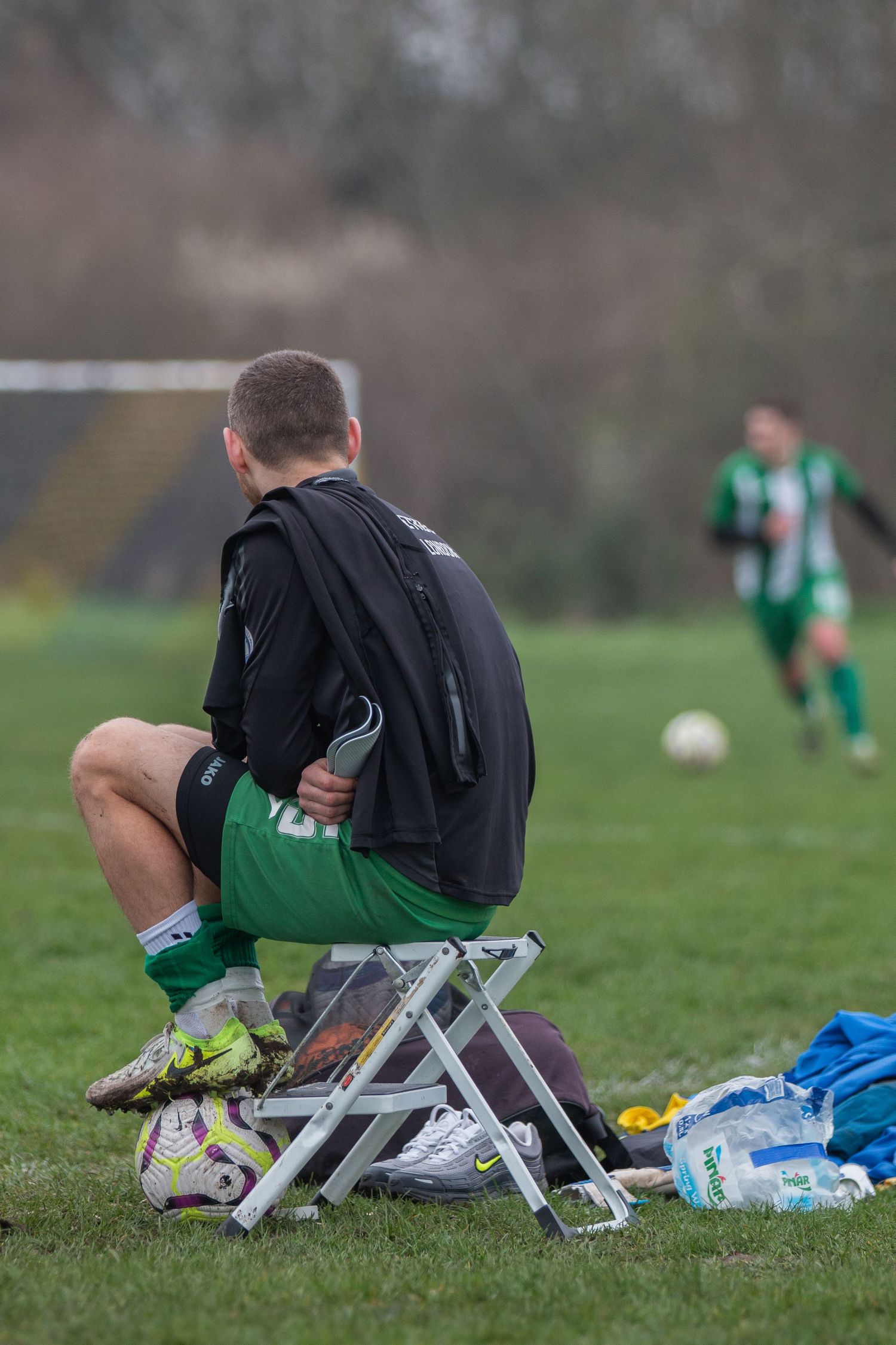 Soccer player resting on a small folding chair on grass field during game break with gear and water bottles nearby.