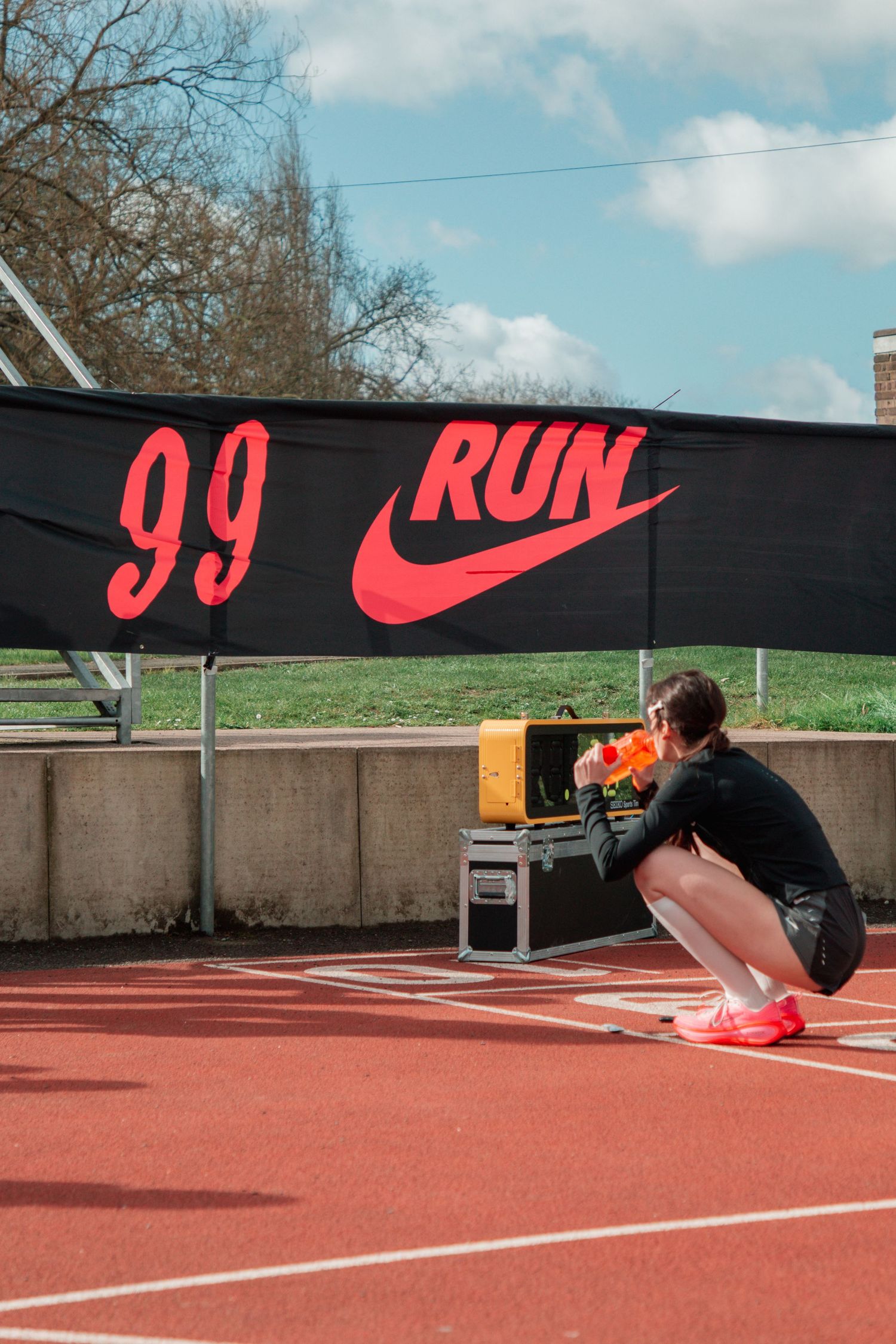 Athlete crouching on a red running track beside a Nike '99 RUN' banner on a sunny day.