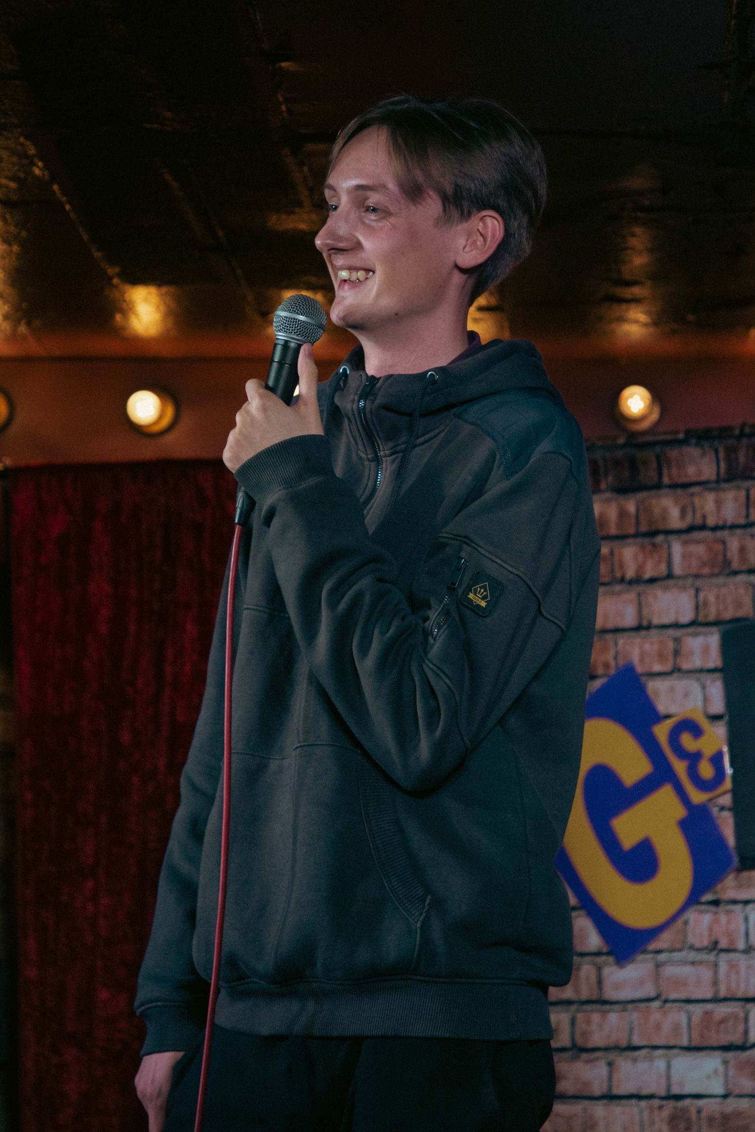 Performer in black hoodie speaking into microphone on brick wall stage.