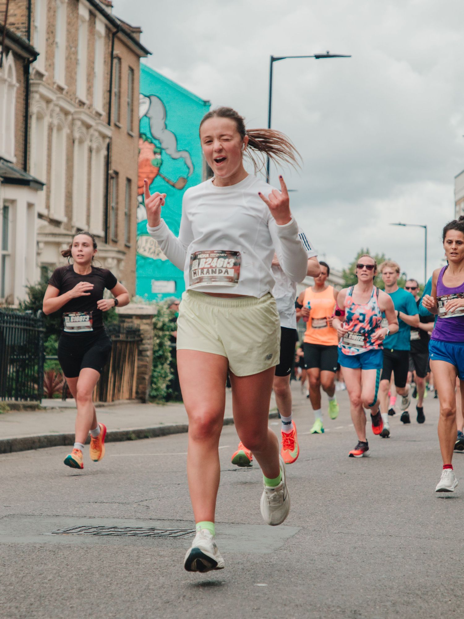 A smiling woman in white top and yellow shorts runs a street marathon, making a rock sign, with other runners behind her.