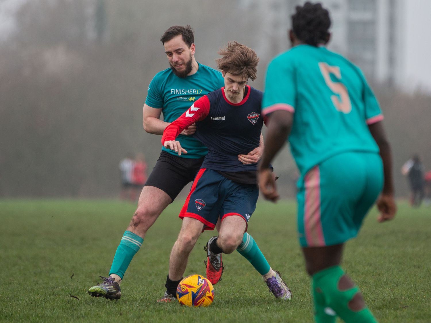 Soccer players compete for possession of the orange ball during a match on a foggy grass field.