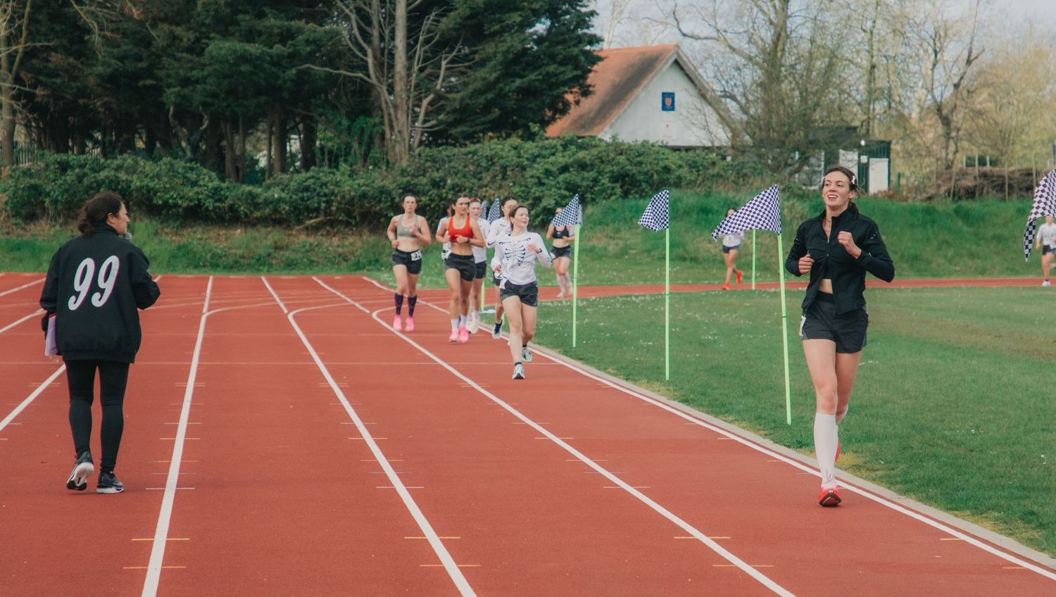 Athletes racing on a red outdoor track during a competitive running event, with spectators and trees in the background.