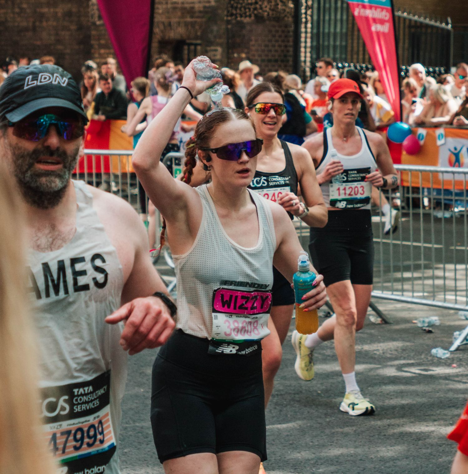 Runners competing in a city marathon, wearing race bibs, including a woman in purple sunglasses and a man with James on his shirt.