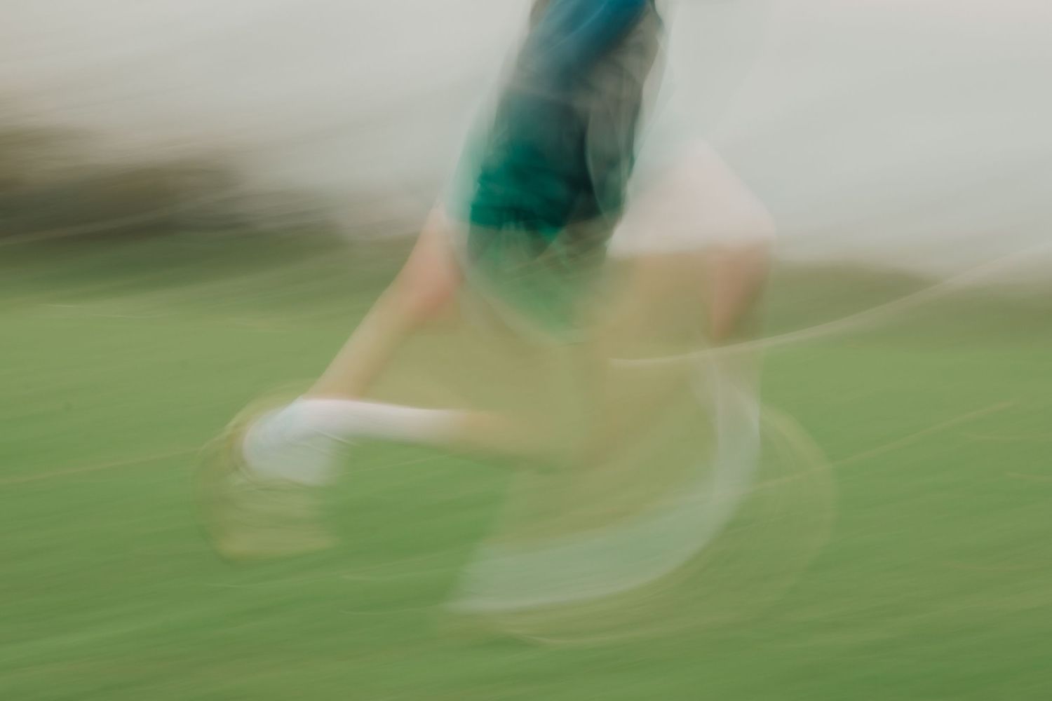 Blurry motion shot of a person running on green grass, holding a white frisbee, wearing teal shorts.