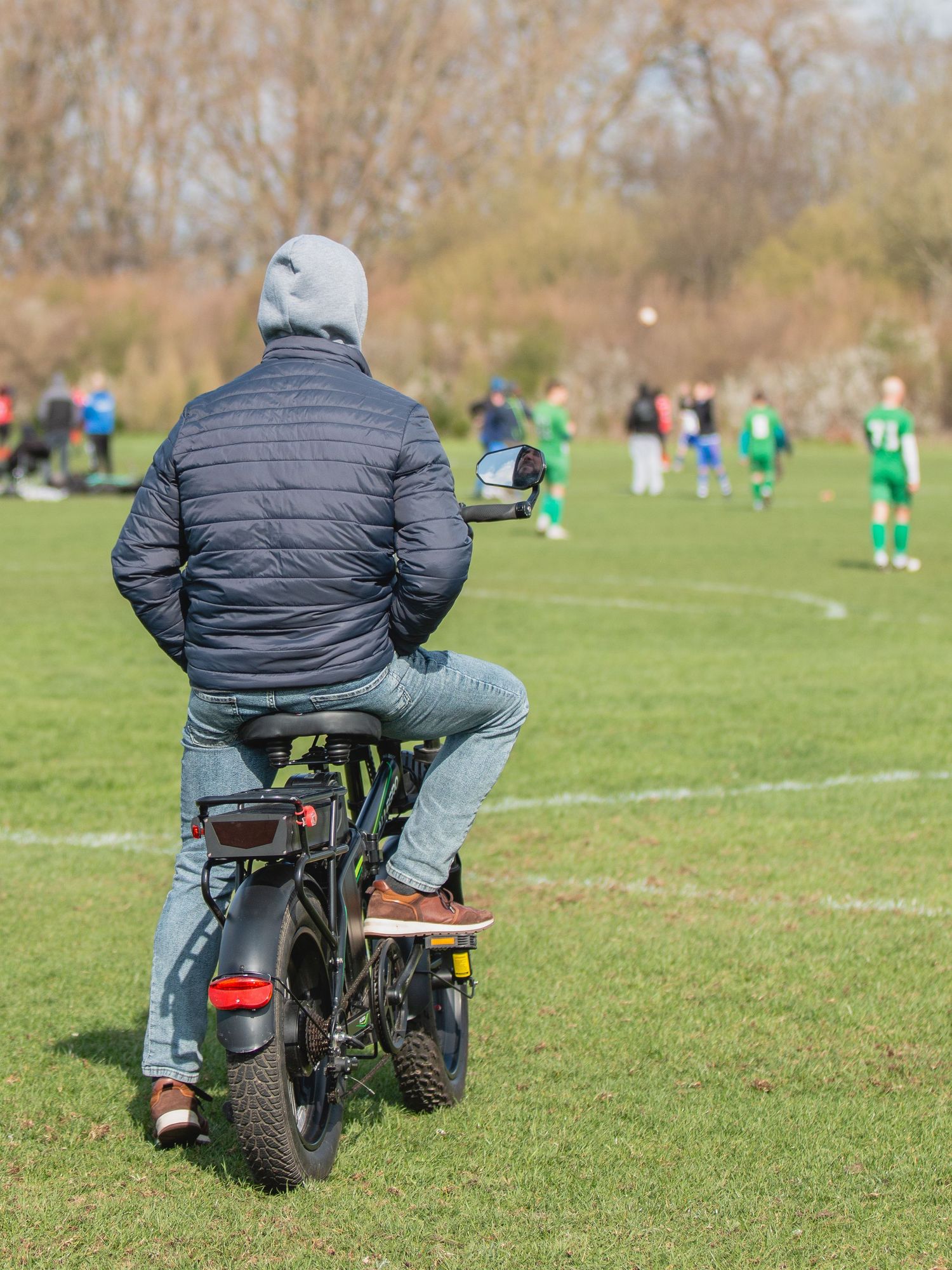 Person in black jacket sits on motorcycle watching youth soccer match on grassy field during fall season.