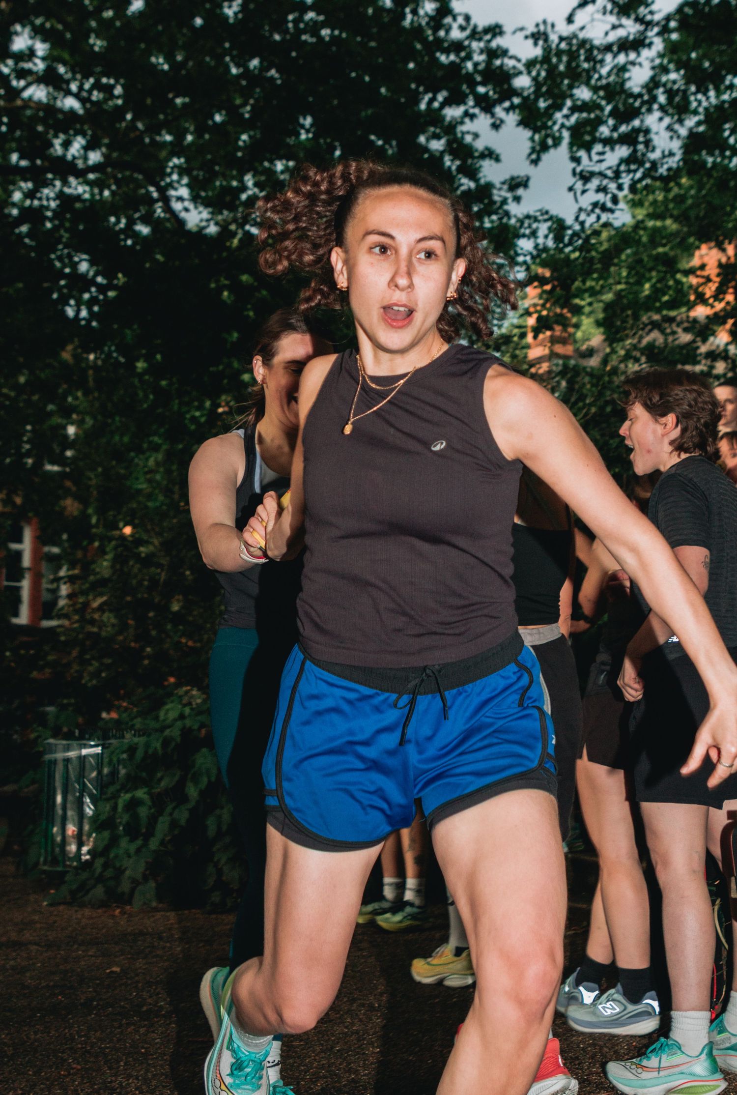 Woman in blue shorts and black tank top running outdoors with other runners and green trees in background.