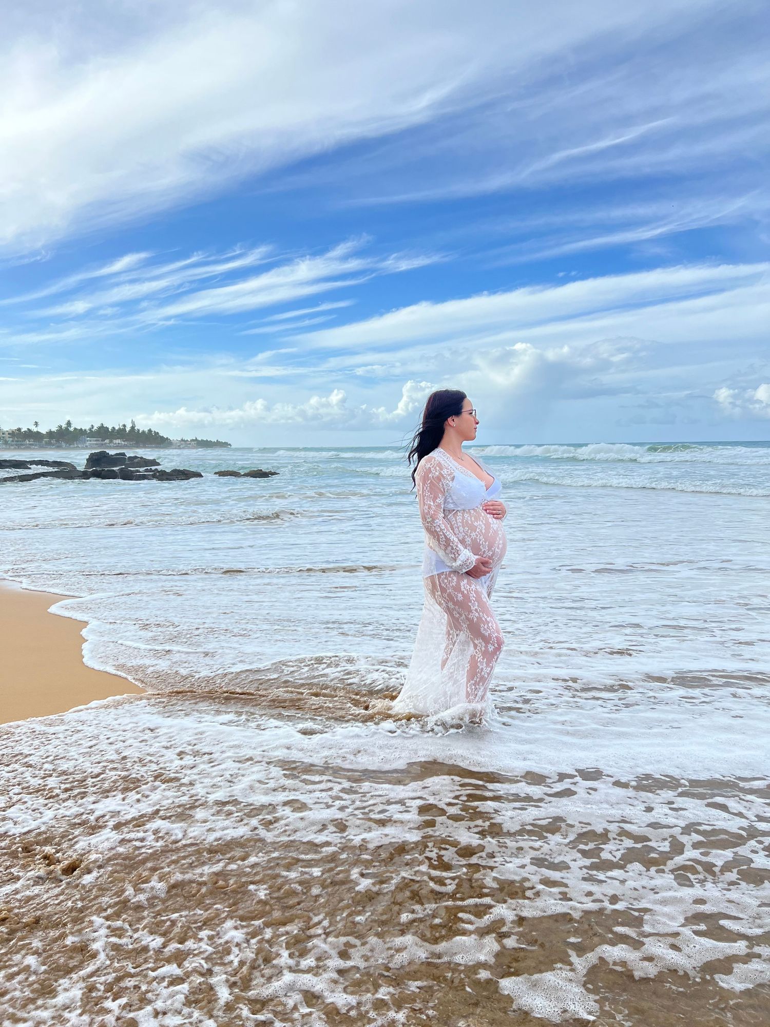 Figure in sheer dress stands in ocean waves at beach during sunset.