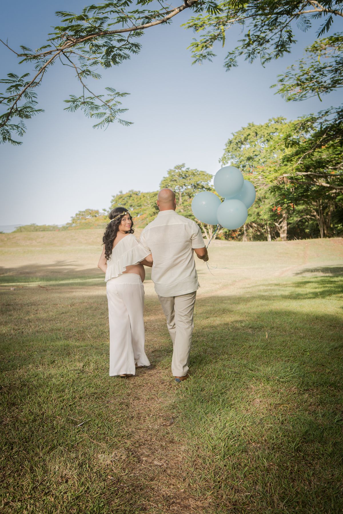 A couple walks on a grassy path holding blue balloons against a sunny outdoor backdrop.