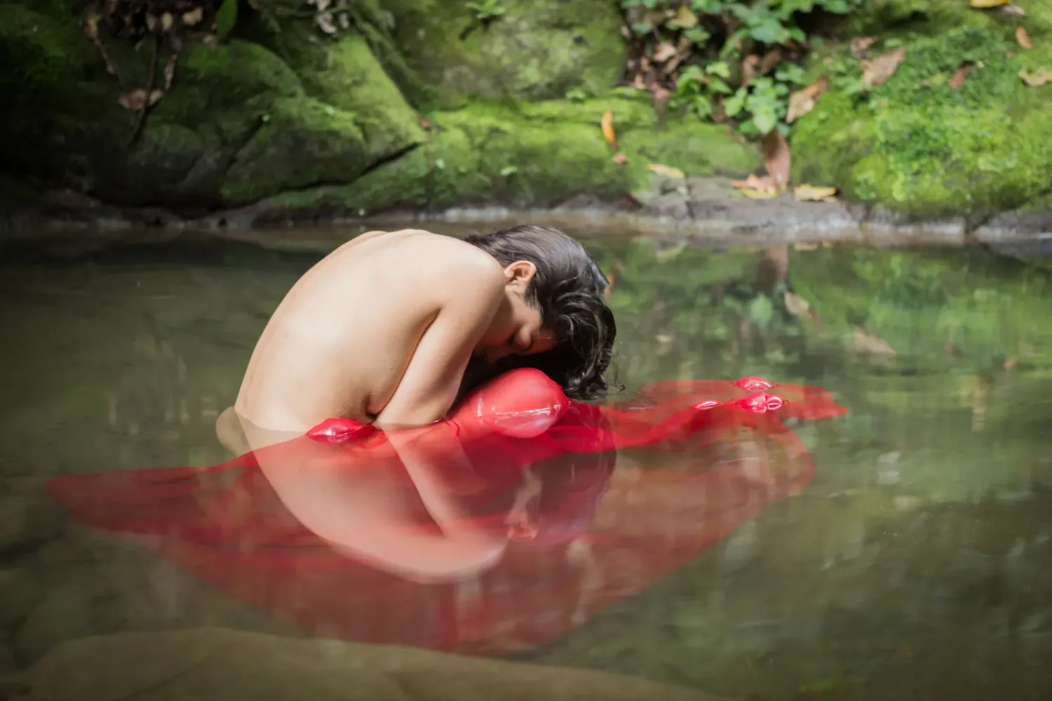 Figure in red fabric reflected in still water surrounded by green foliage.