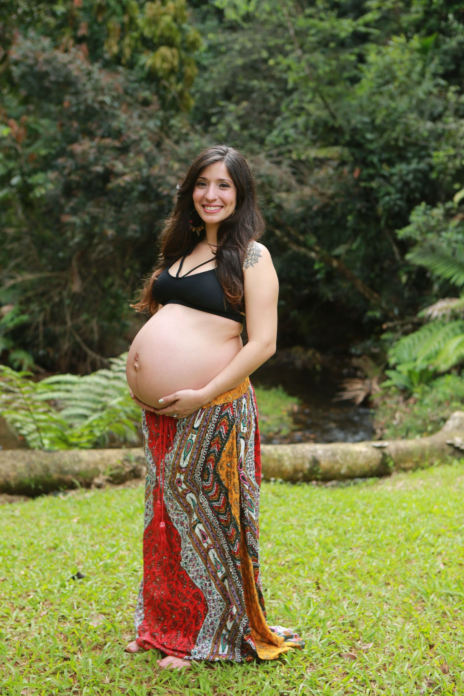 Expectant mother in a colorful patterned skirt poses outdoors in a garden setting.