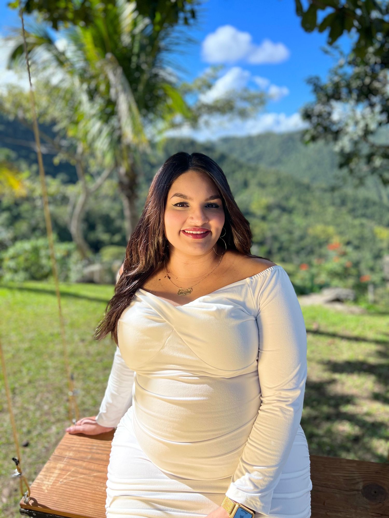 Woman in white dress poses outdoors with tropical mountain landscape behind.