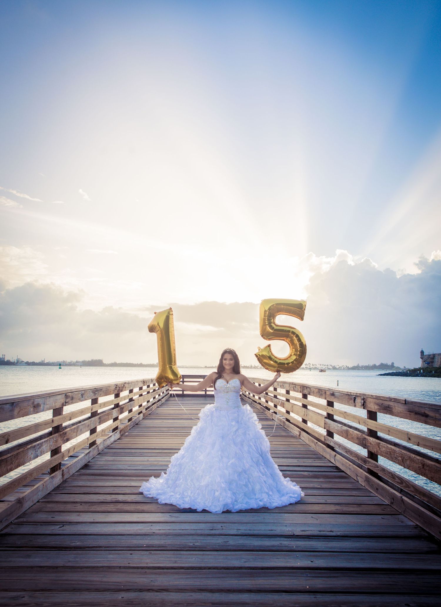 Quinceañera photo with gold number 15 balloons on wooden pier at sunset.