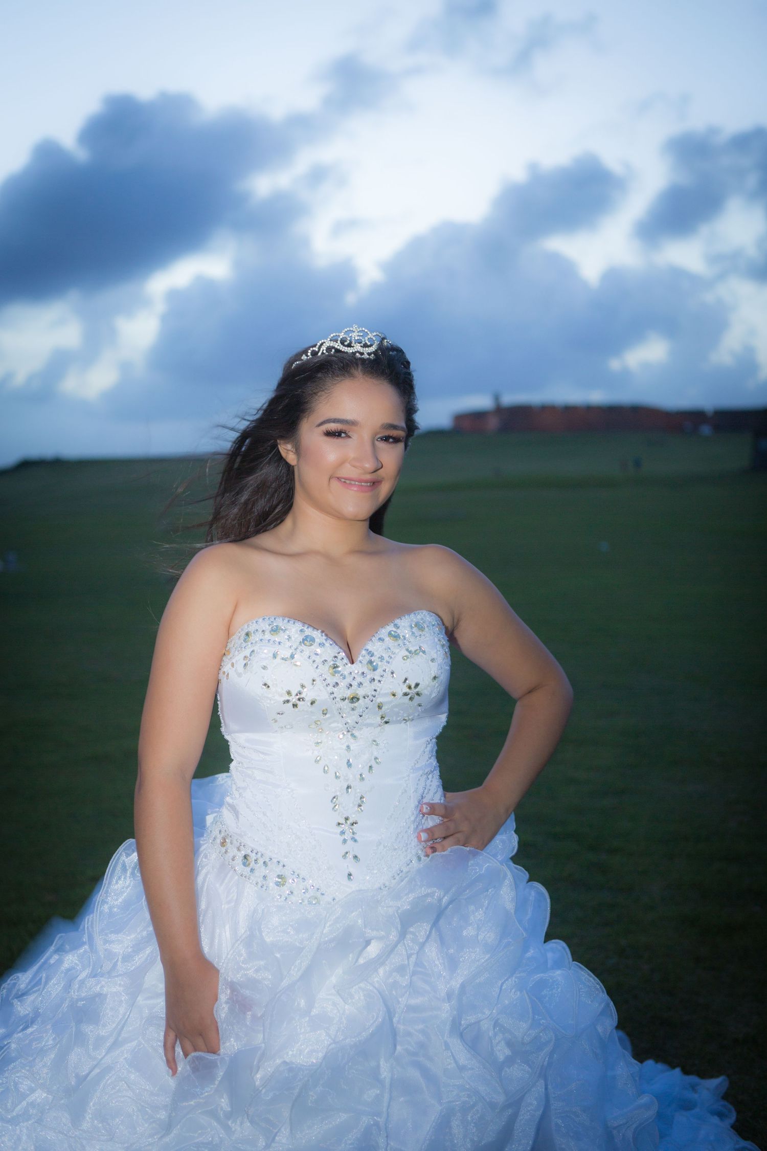 Formal portrait in white ballgown dress with tiara against blue sky background.