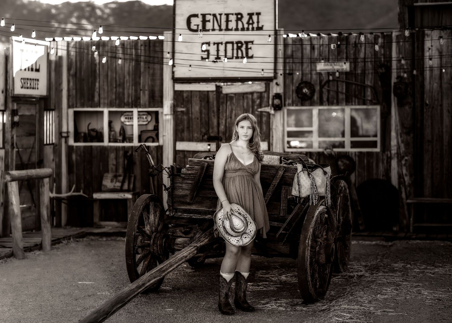 Black and white portrait of young woman in dress with cowboy hat by General Store wagon.