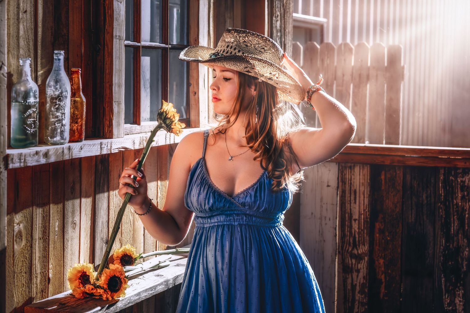 Young woman in blue dress and cowboy hat holding sunflower in sunlit rustic barn interior.