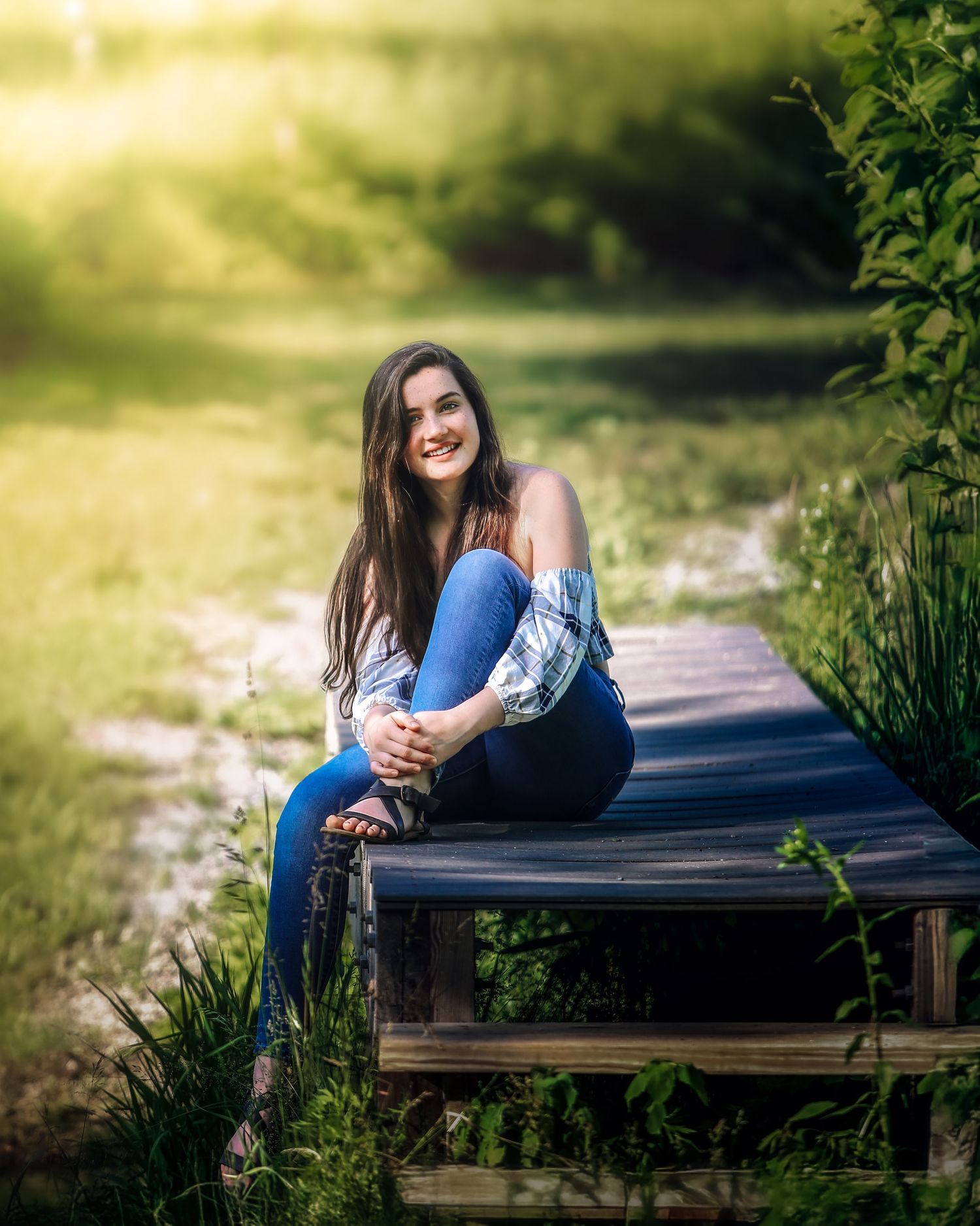 Smiling teenage girl in jeans sitting on wooden bridge surrounded by green nature.