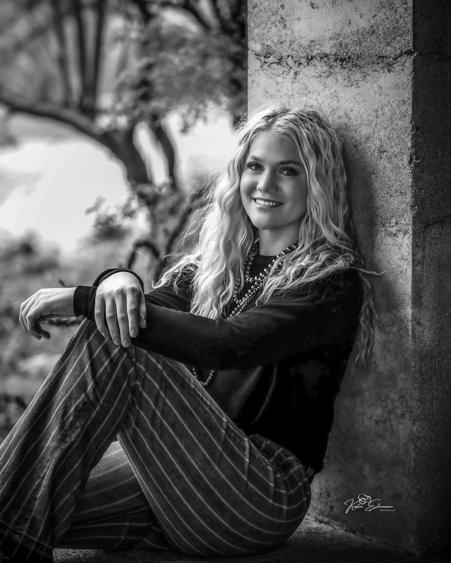 Black and white portrait of smiling blonde teen girl leaning against stone wall in striped pants.