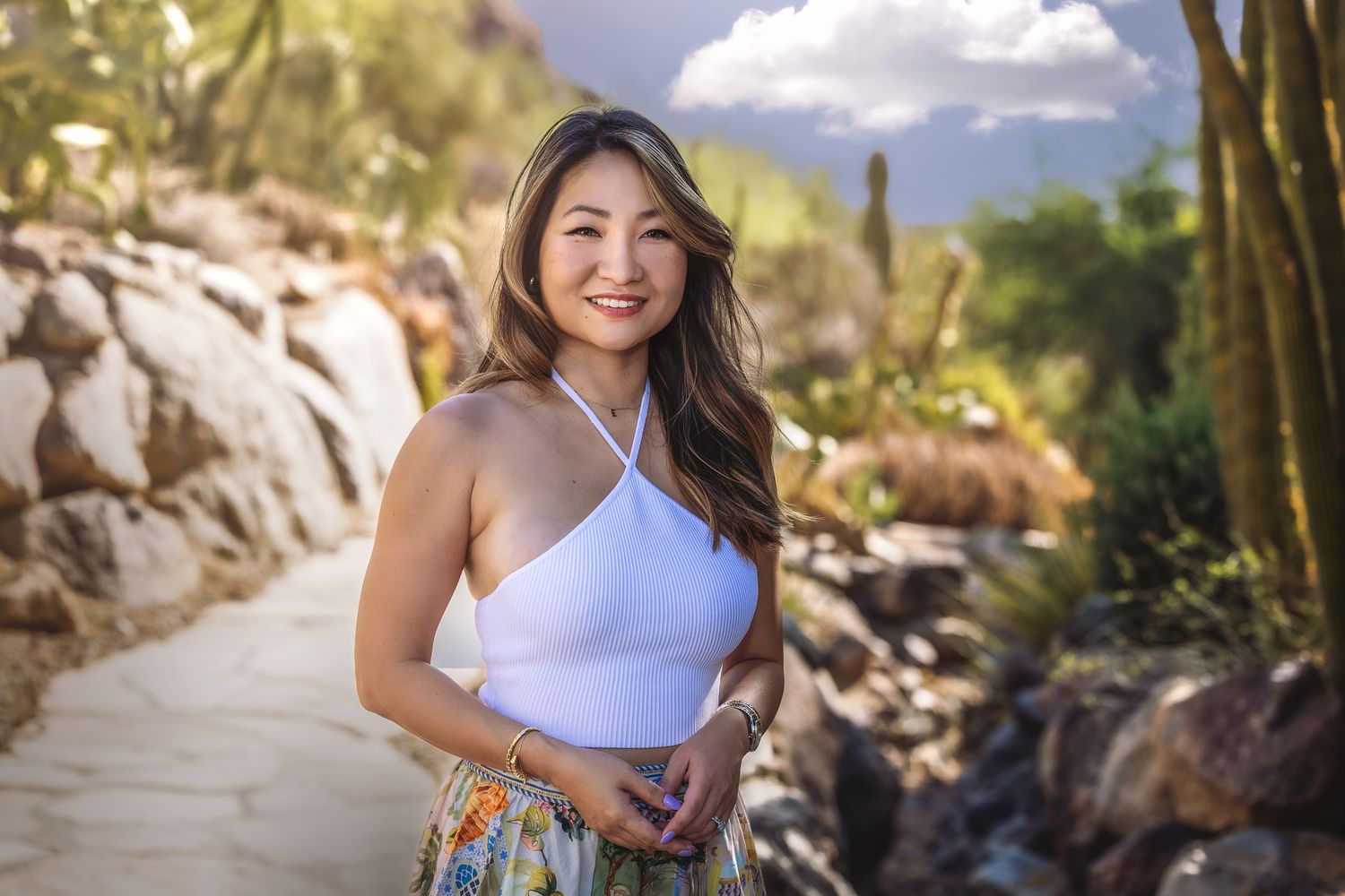 Young woman in a white halter top and floral skirt smiling on a desert garden path with cacti.