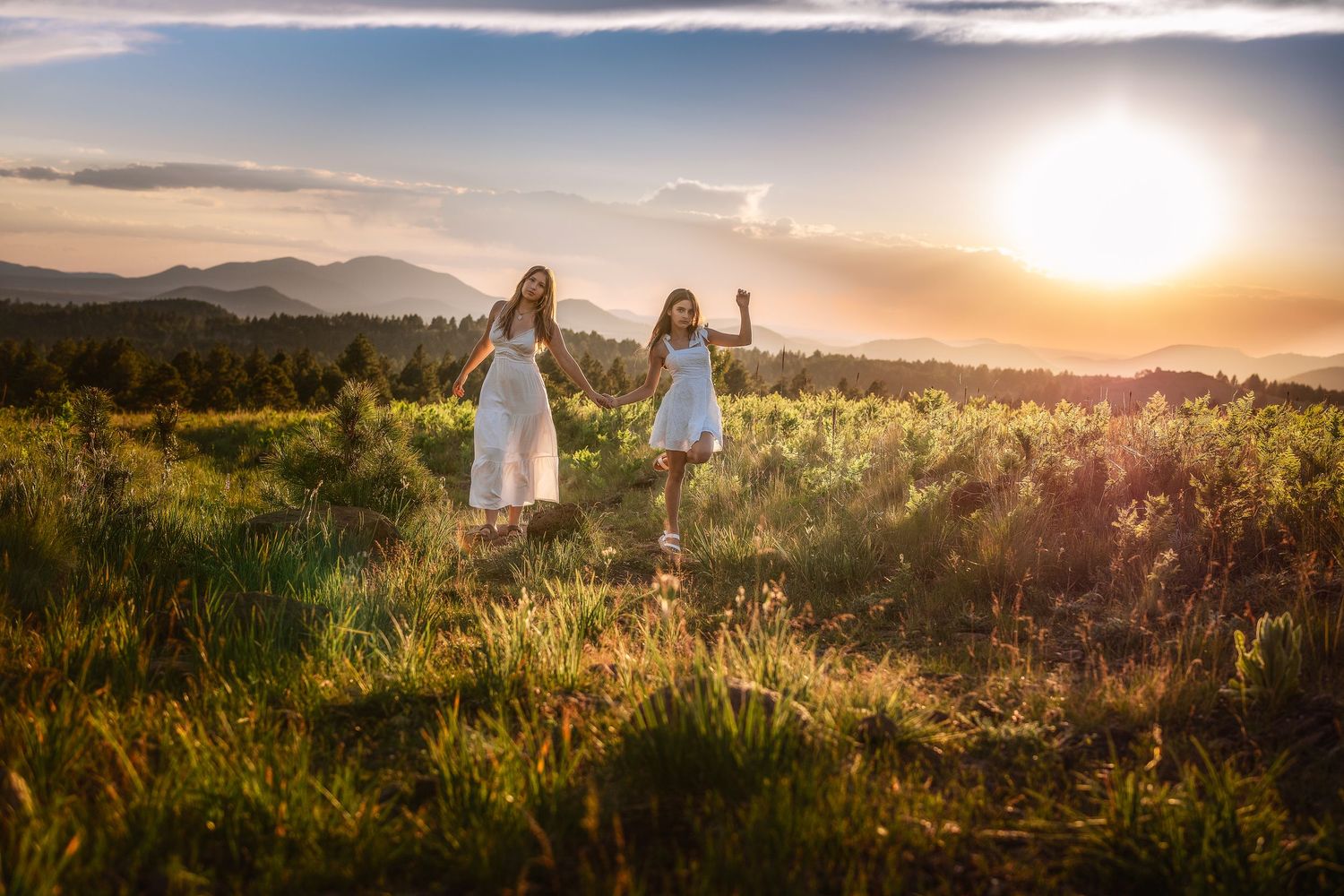 Two women in white dresses stand in a sunlit meadow with mountains and a golden sunset in the background.