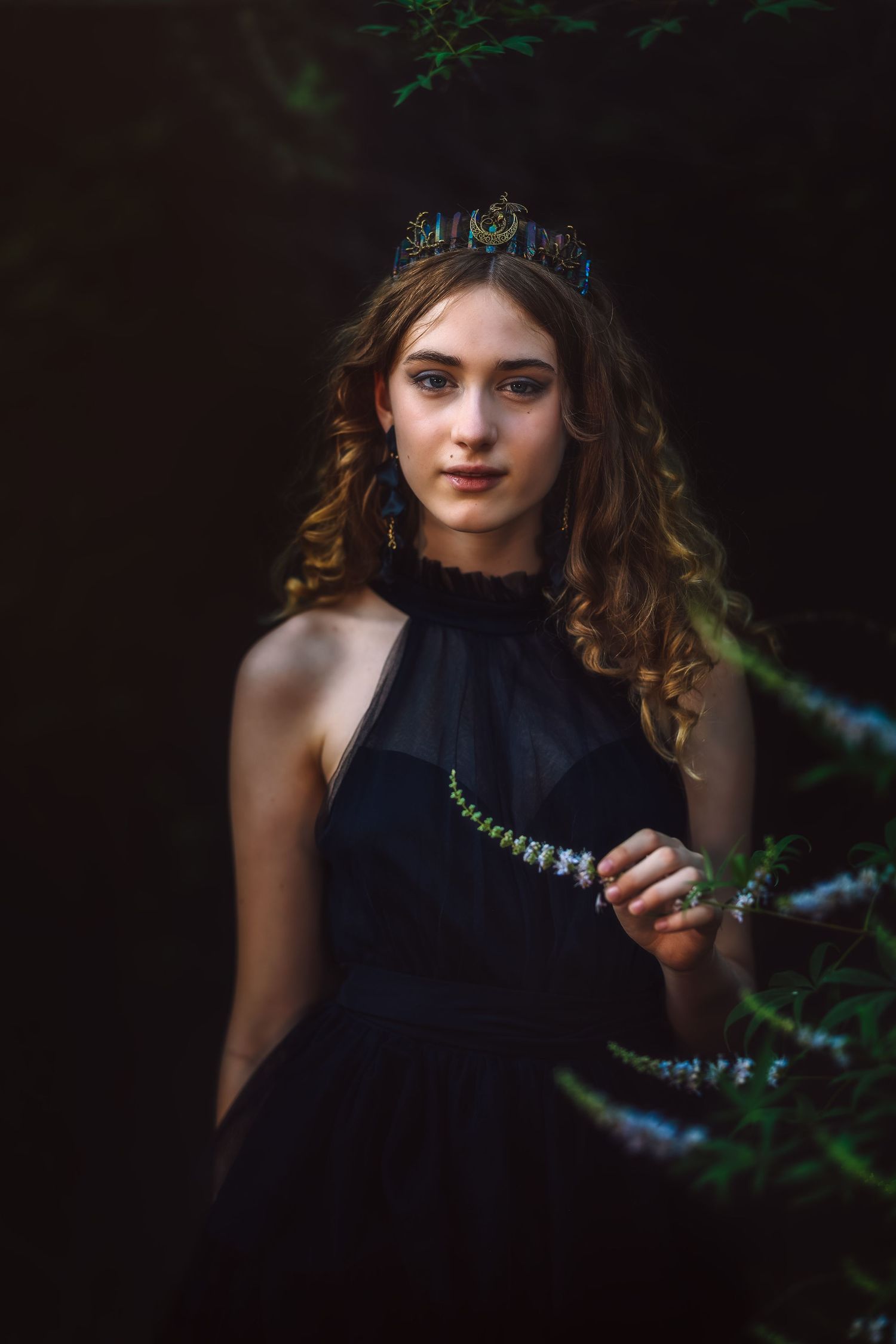 Young woman in black halter dress and decorative crown poses among dark forest greenery.