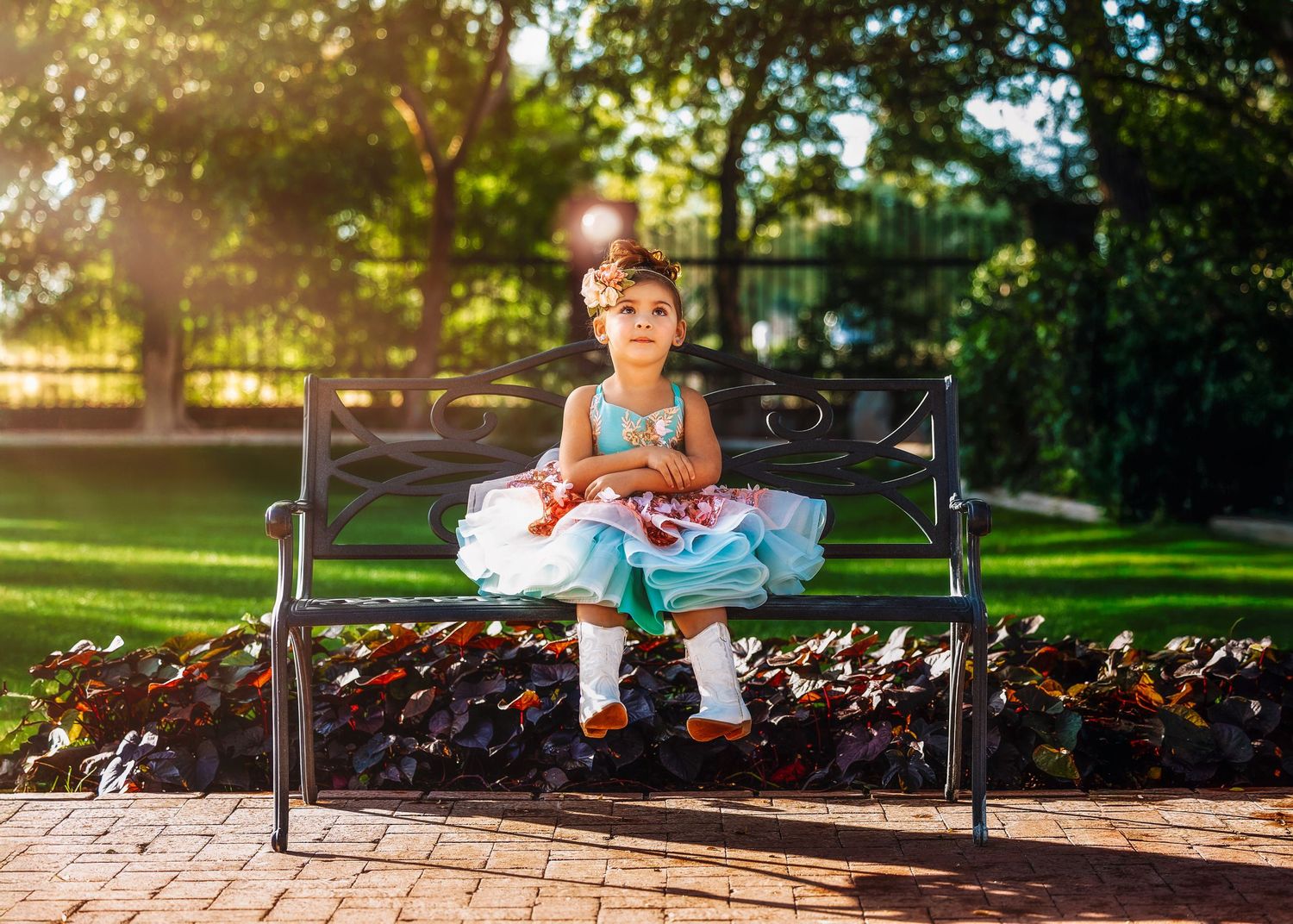 A little girl in a colorful ruffled dress sits on a park bench holding an ice cream cone outdoors.