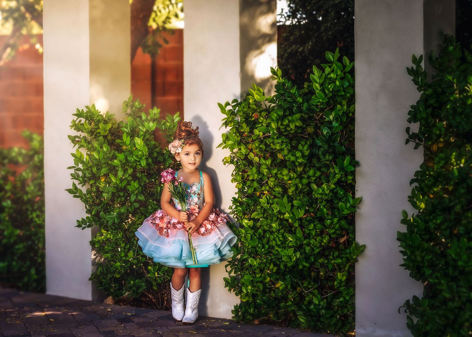 A young girl in a layered teal and pink dress with white boots stands between green bushes in sunlight.