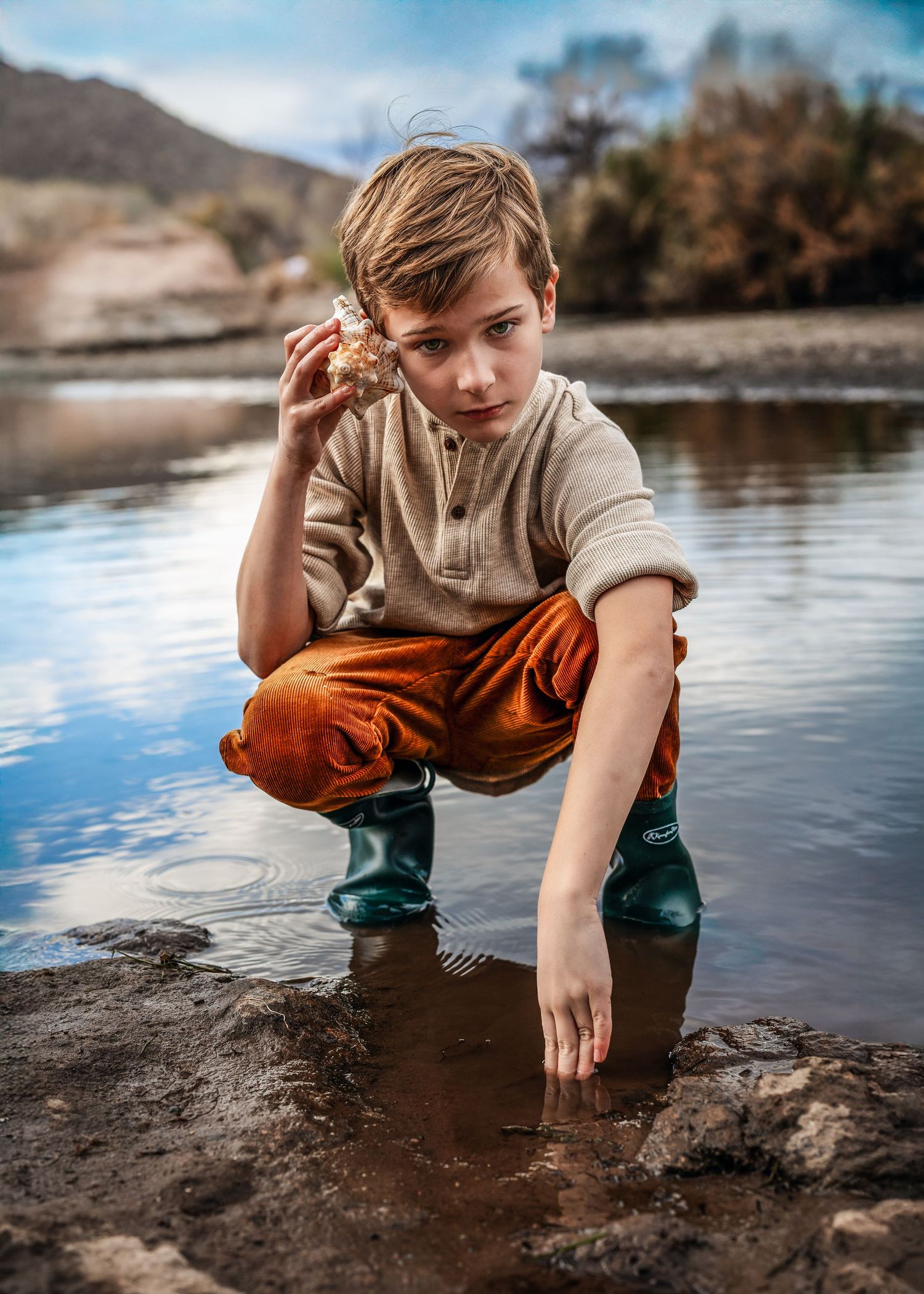 Boy squatting in shallow water holding a rock while touching the water surface.