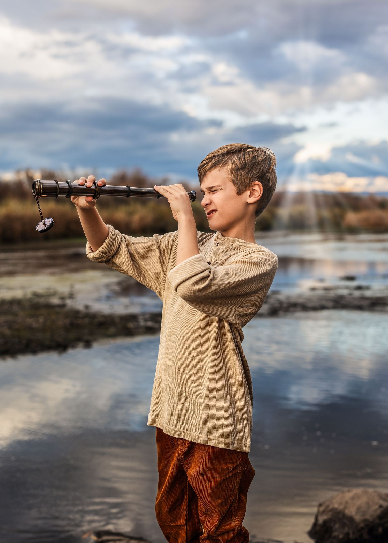 Young boy in a linen shirt peers through a vintage telescope beside a reflective lake under a cloudy sky.