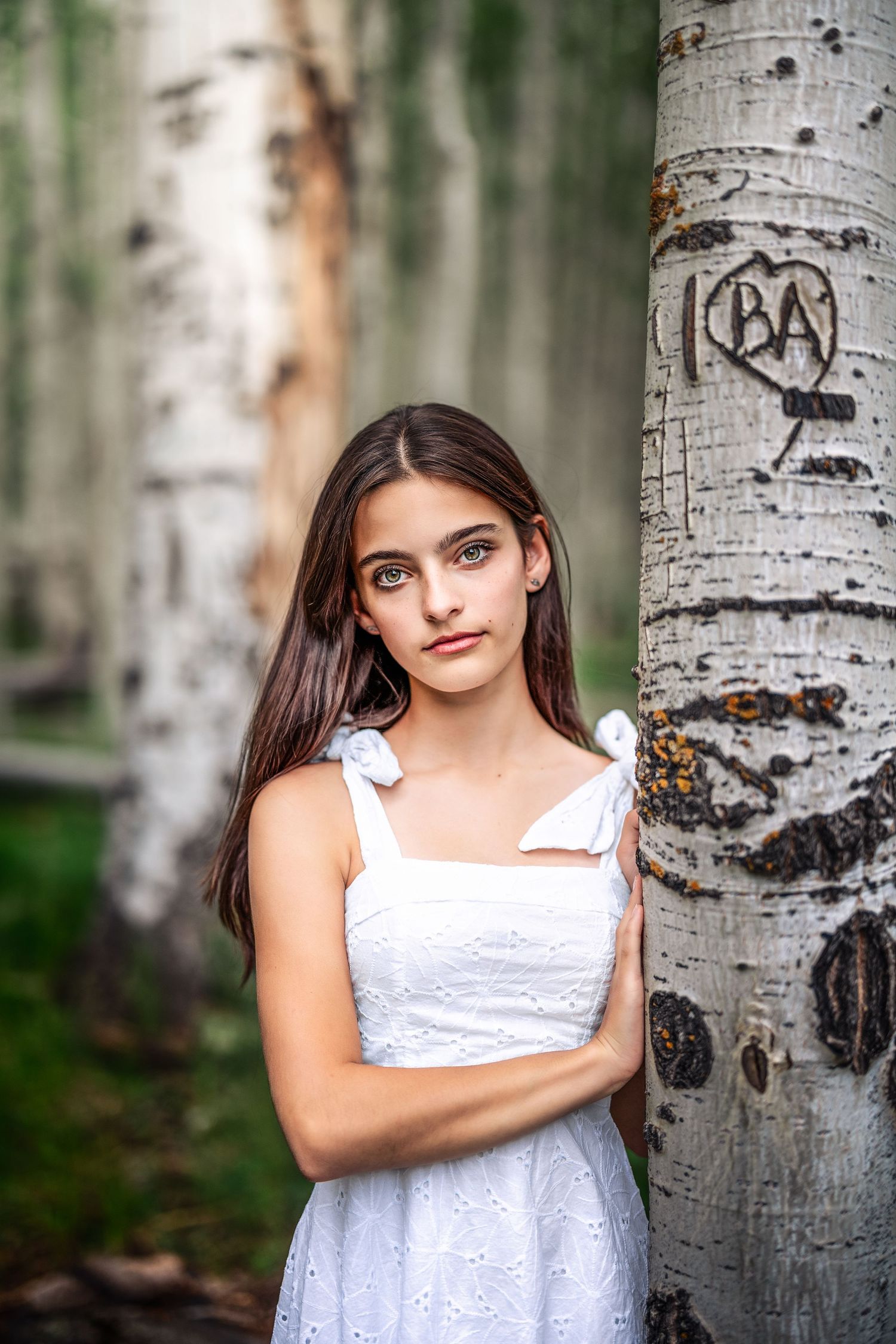 Young brunette girl in white sundress leaning against birch tree with carved initials in forest setting.