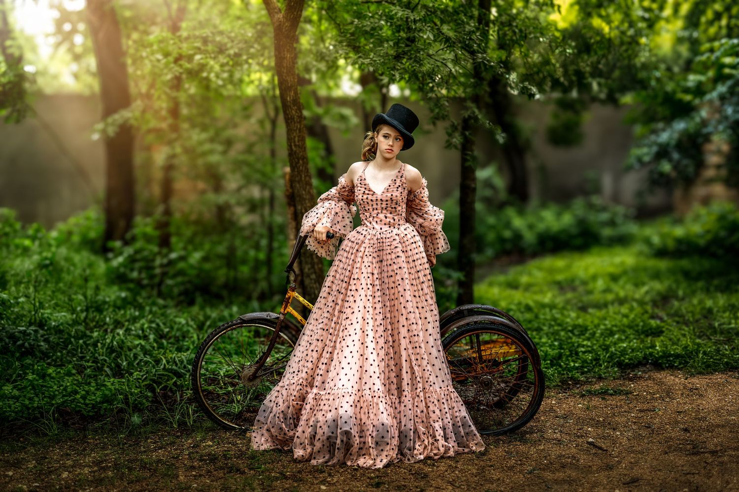Woman in vintage polka dot gown and black hat poses beside a bicycle in a sunlit green forest.