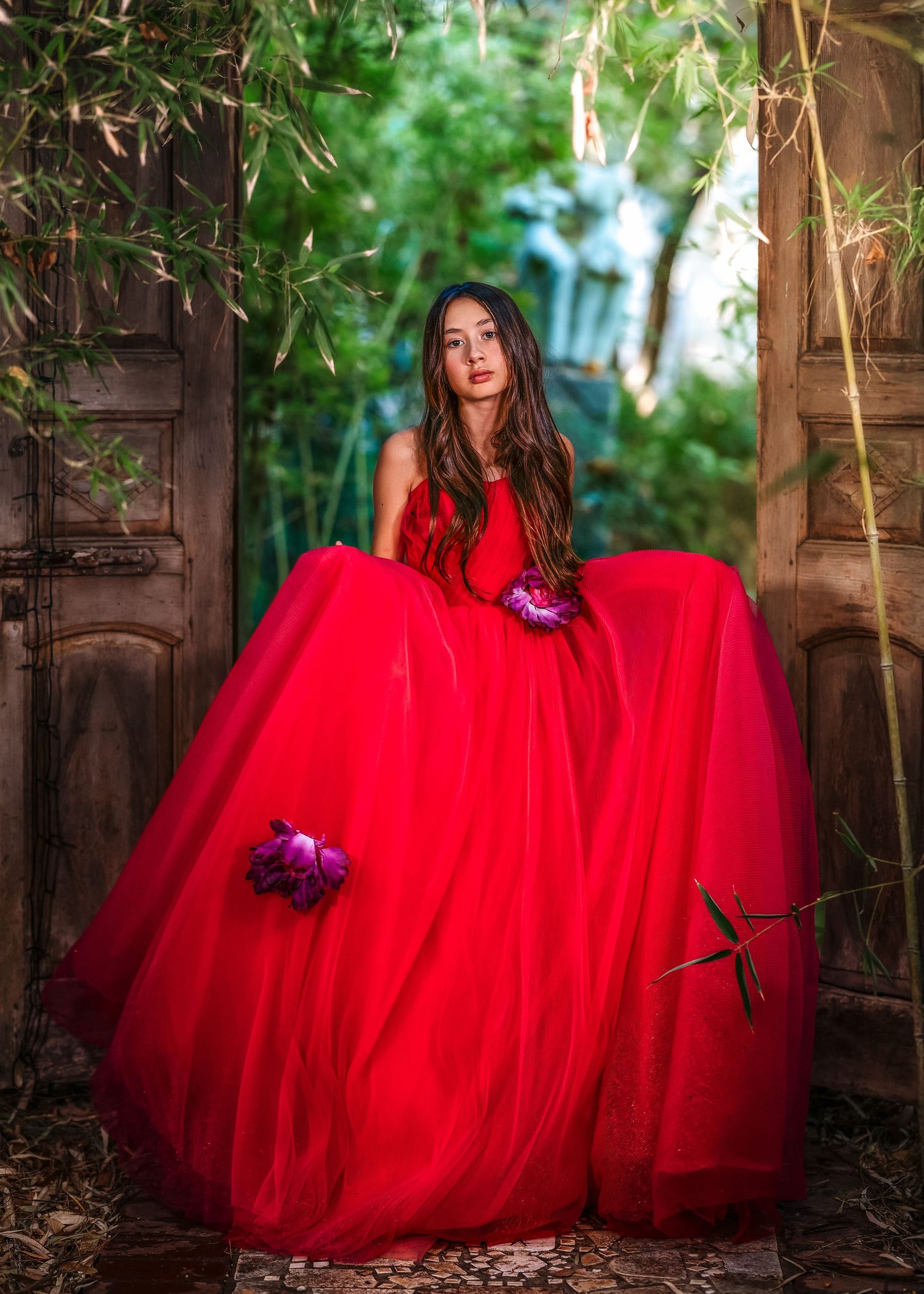 Young woman in a voluminous red ball gown standing in a rustic doorway surrounded by lush greenery.