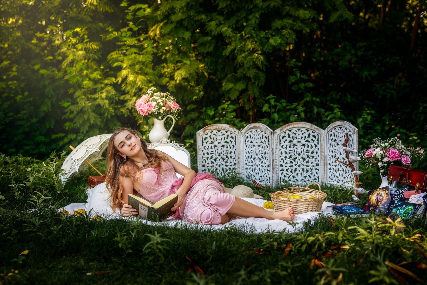 Woman in pink dress reading book on white ornate bench in sunlit garden with parasol and flowers.