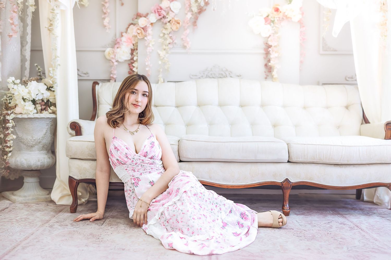 Young woman in pink floral dress sitting elegantly on floor beside white vintage sofa with flowers.