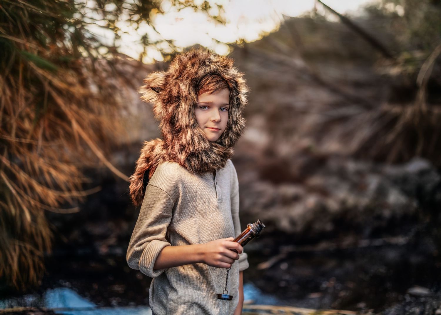A child wearing a furry hooded hat holds a stick and stands among trees in warm golden outdoor light.