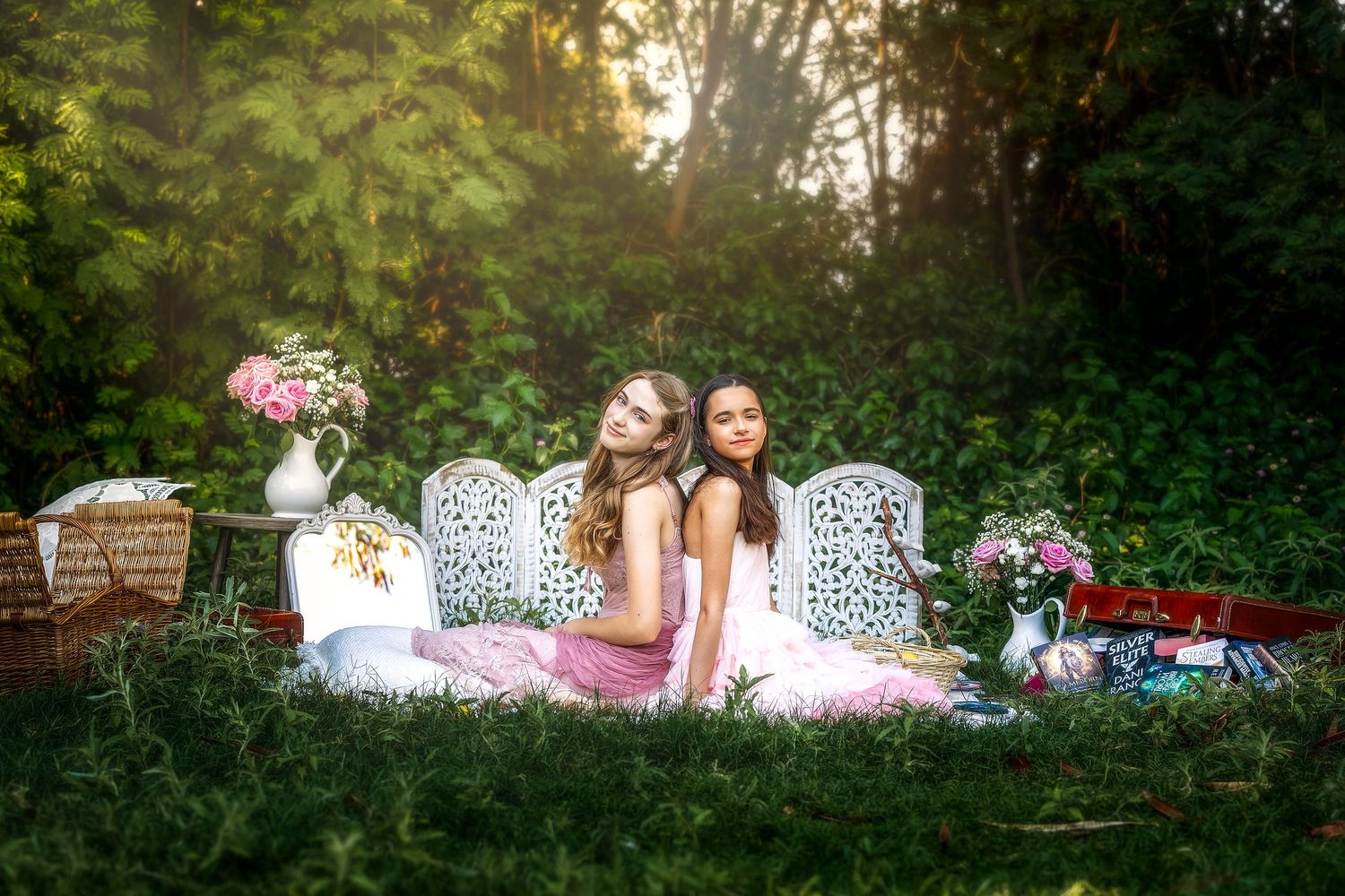 Two young women in floral dresses sit back-to-back on a white ornate bench surrounded by lush greenery and flowers.