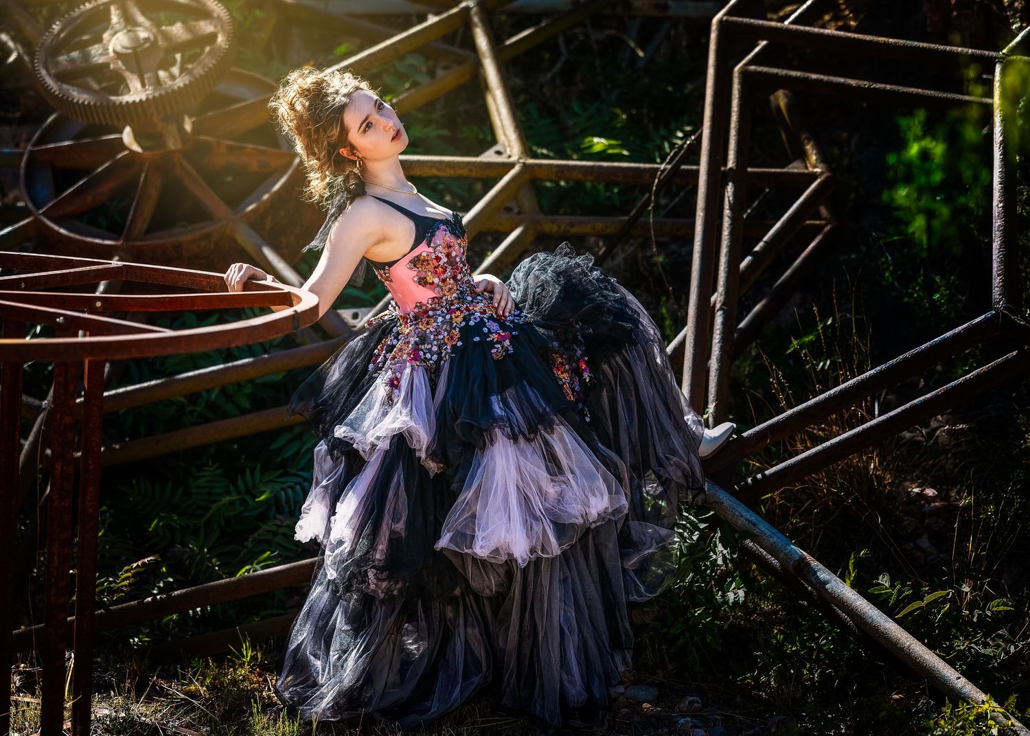 Woman in ornate dark floral gown poses dramatically among rustic metal structures outdoors in dappled sunlight.