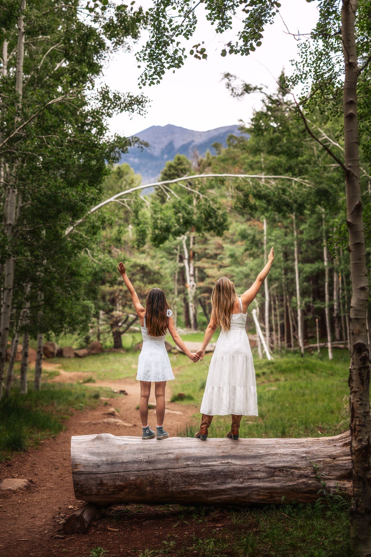 Two girls in white dresses stand on a log holding hands with arms raised in a lush forest with mountain views.