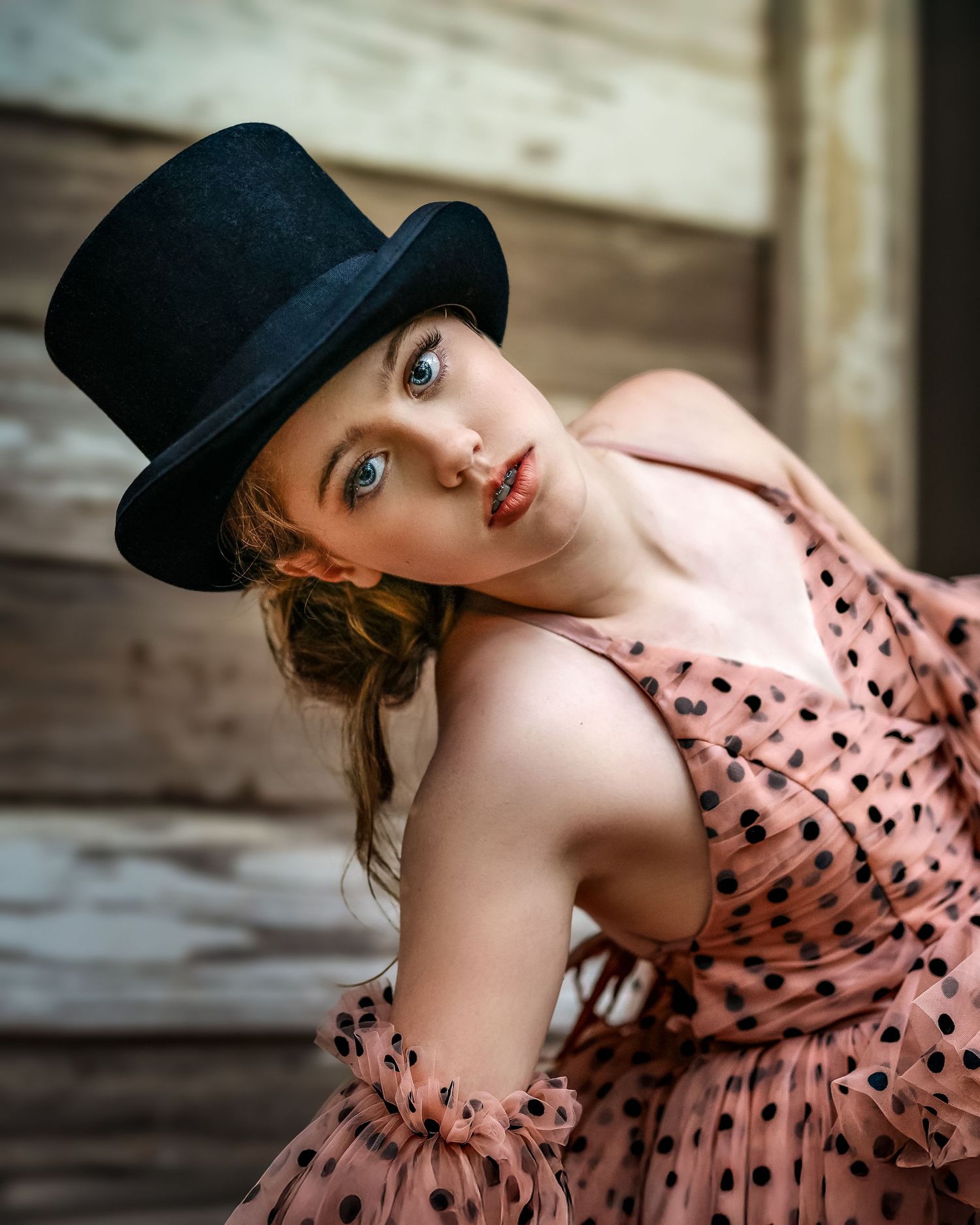 Young woman in pink polka dot dress and black top hat posing against rustic wooden wall.