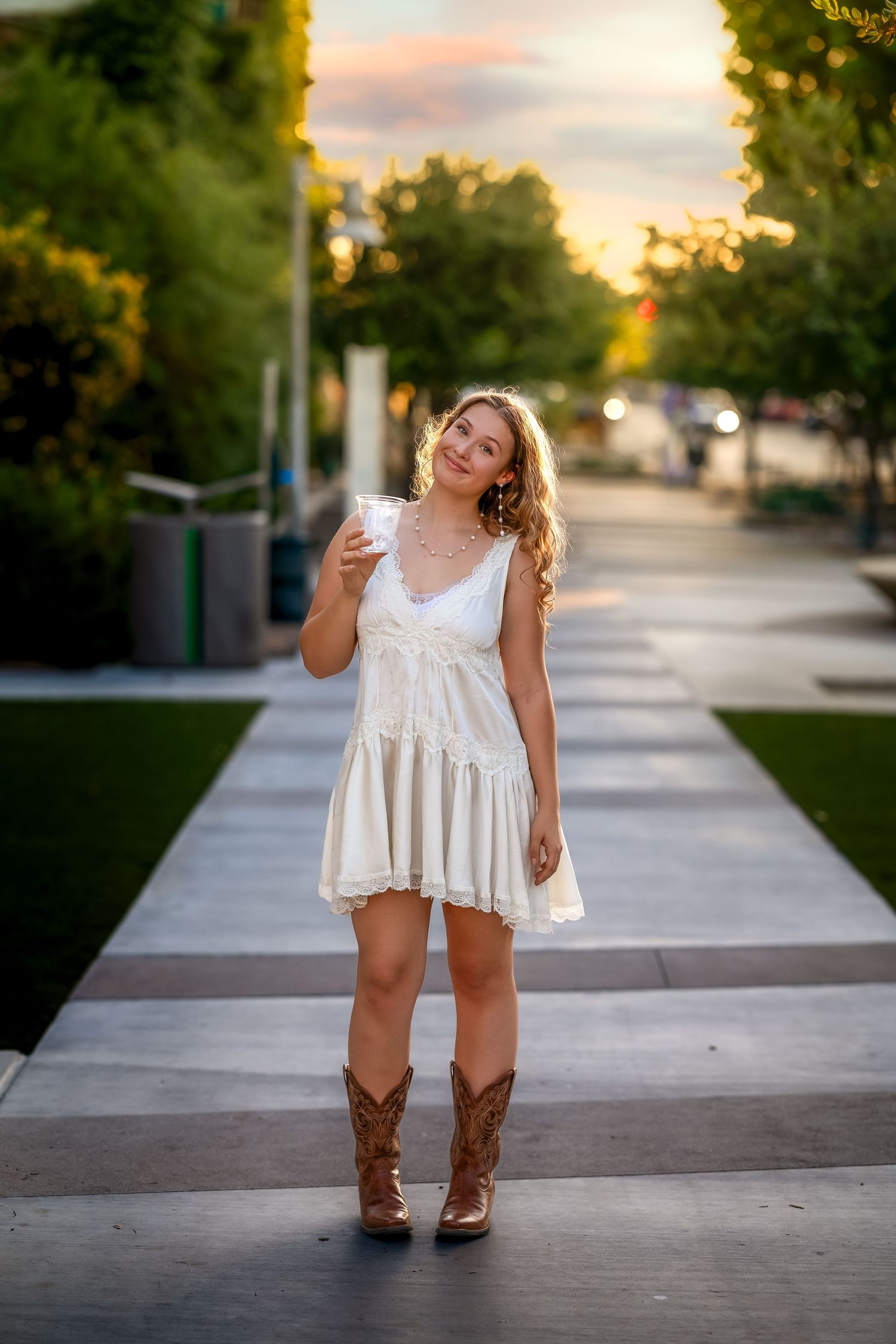 Young woman in white dress and cowboy boots holds a drink outdoors at sunset on a tree-lined walkway.