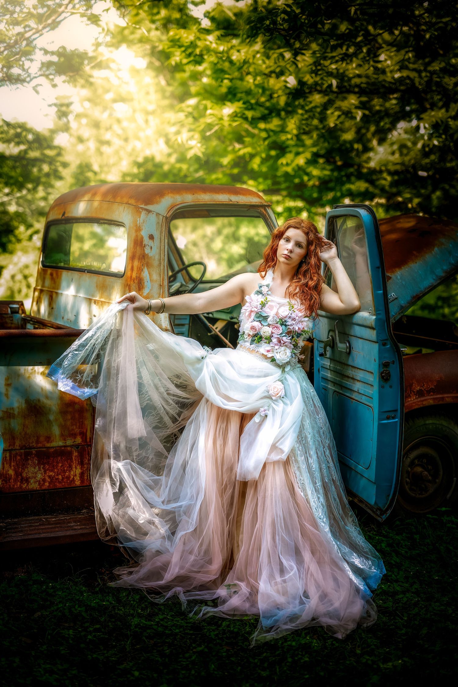 A red-haired woman in a floral tulle gown poses beside a rusty vintage blue truck in a lush green outdoor setting.