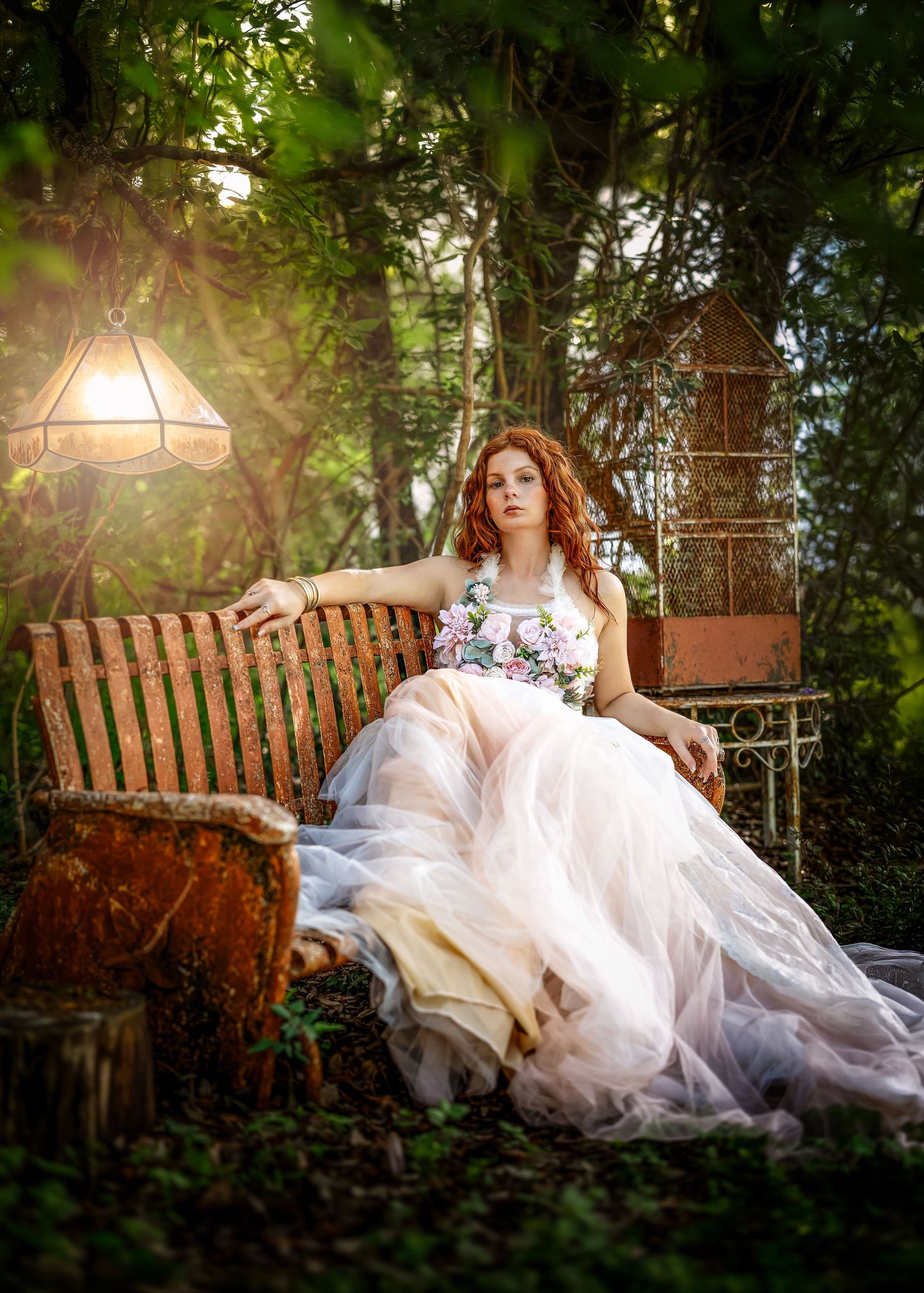 Red-haired woman in floral tulle gown sits on rustic bench in enchanted garden with vintage lamp and birdcage.
